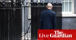 英国财政大臣约翰·希利在唐宁街手持预算文件箱，背景呈现英国生活成本与预算争议